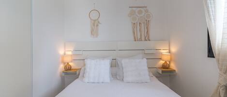 A neatly made bed with white linens, two bedside tables with lamps, and wall decorations including a dreamcatcher and a hanging ornament.