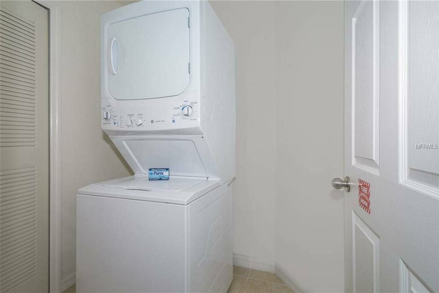 A laundry room with a stacked washer and dryer, a fire exit sign, and a door with 'MFRMLS' label.