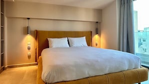 Ein großes Bett mit Kopfteil, zwei Nachttischlampen und Blick auf Gebäude durch ein Fenster.