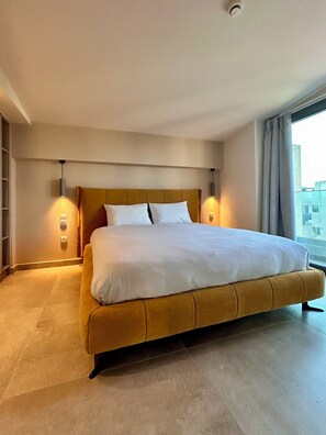 A large bed with a headboard, two bedside lamps, and a view of buildings through a window.