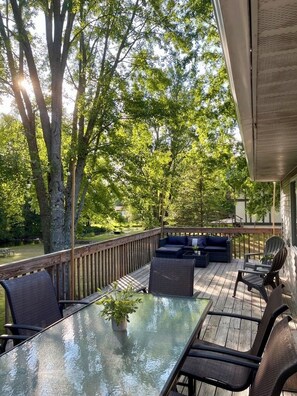 An outdoor deck with a glass table and chairs, surrounded by trees and a wooden railing. - Peaceful Pere Marquette River Retreat (Branch)