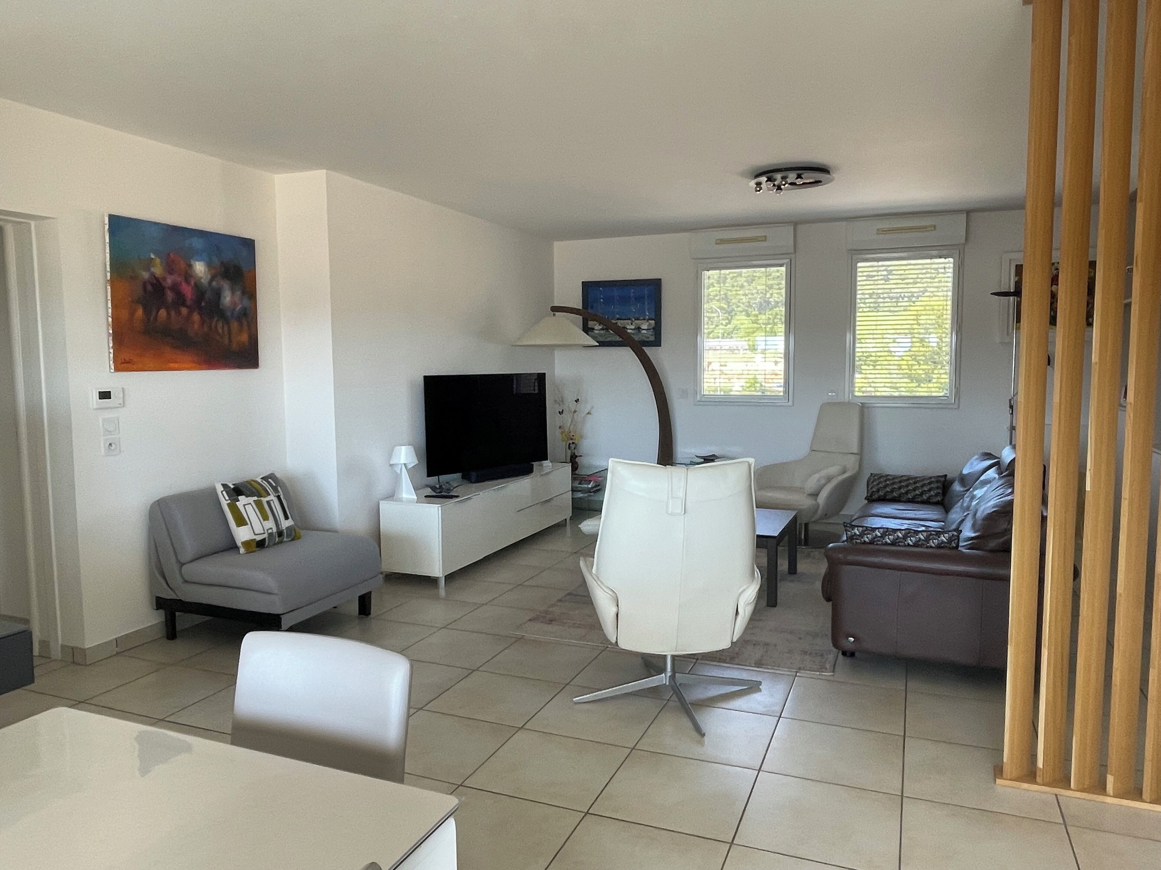Een moderne woonkamer met een flatscreen televisie, een witte bank, een grijze bank, een witte fauteuil en een eettafel.