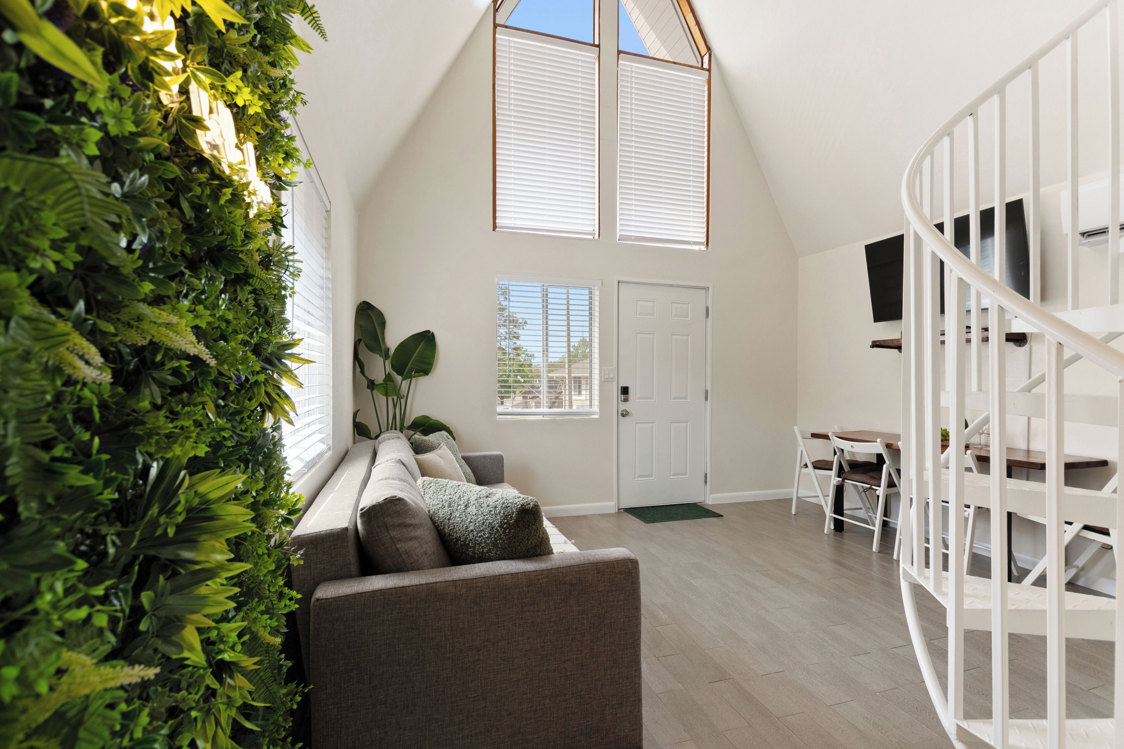 A room with a high ceiling, a built-in plant wall, a gray sofa, a white door, and a white spiral staircase.
