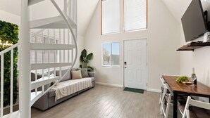 A room with a staircase, a sofa, a dining table with chairs, a TV mounted on the wall, and a door.