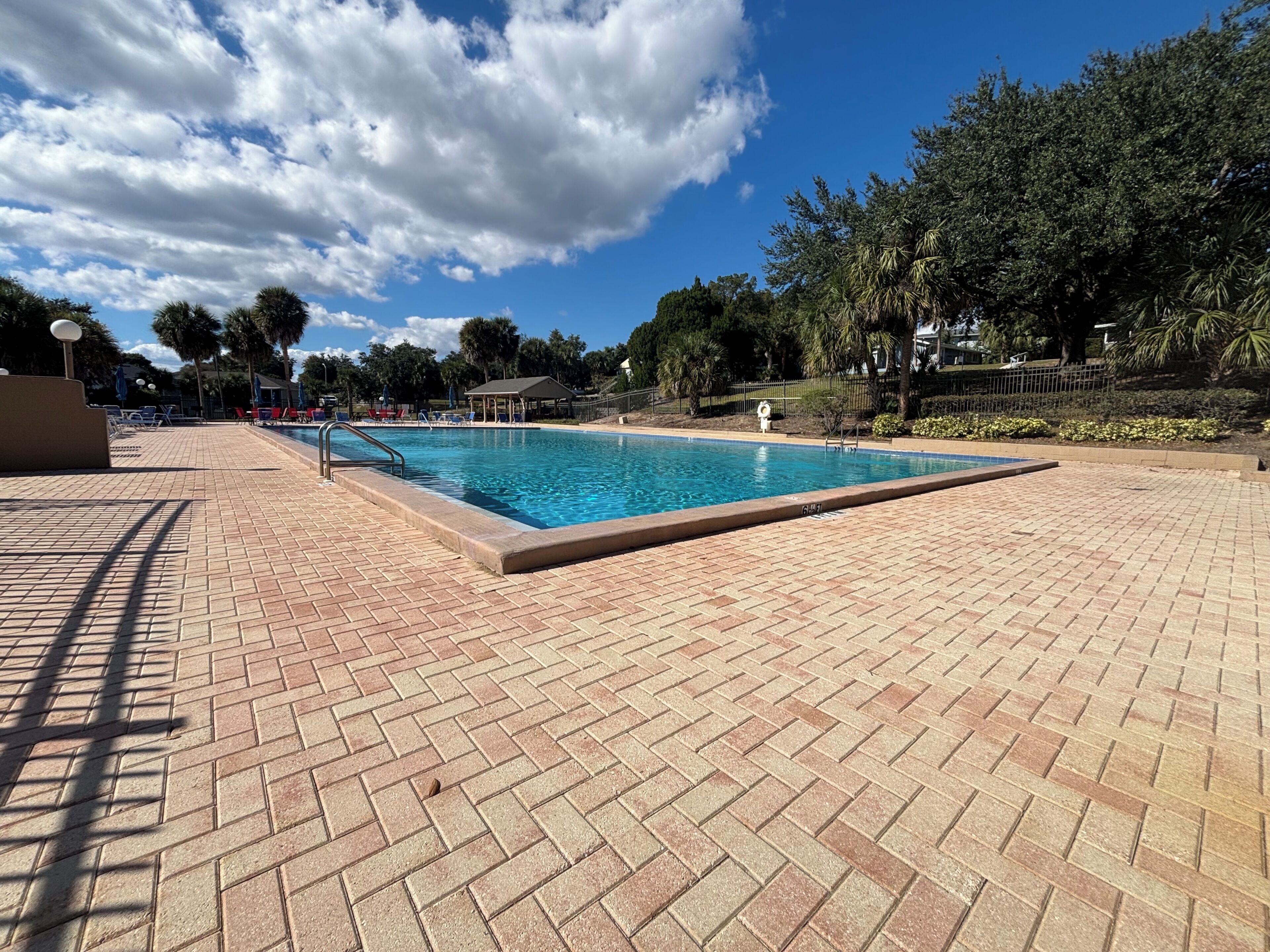 Une piscine entourée d’arbres, avec une terrasse en briques et un ciel dégagé.
