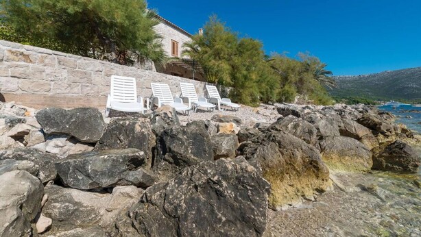 Un mur de pierres, des fauteuils de jardin blancs, un bâtiment avec un toit en tuiles et une côte rocheuse bordée d’une eau claire.