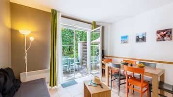 Una habitación con cama, escritorio y zona de comedor con sillas. Hay una ventana con cortinas, una lámpara y una puerta que da al exterior.