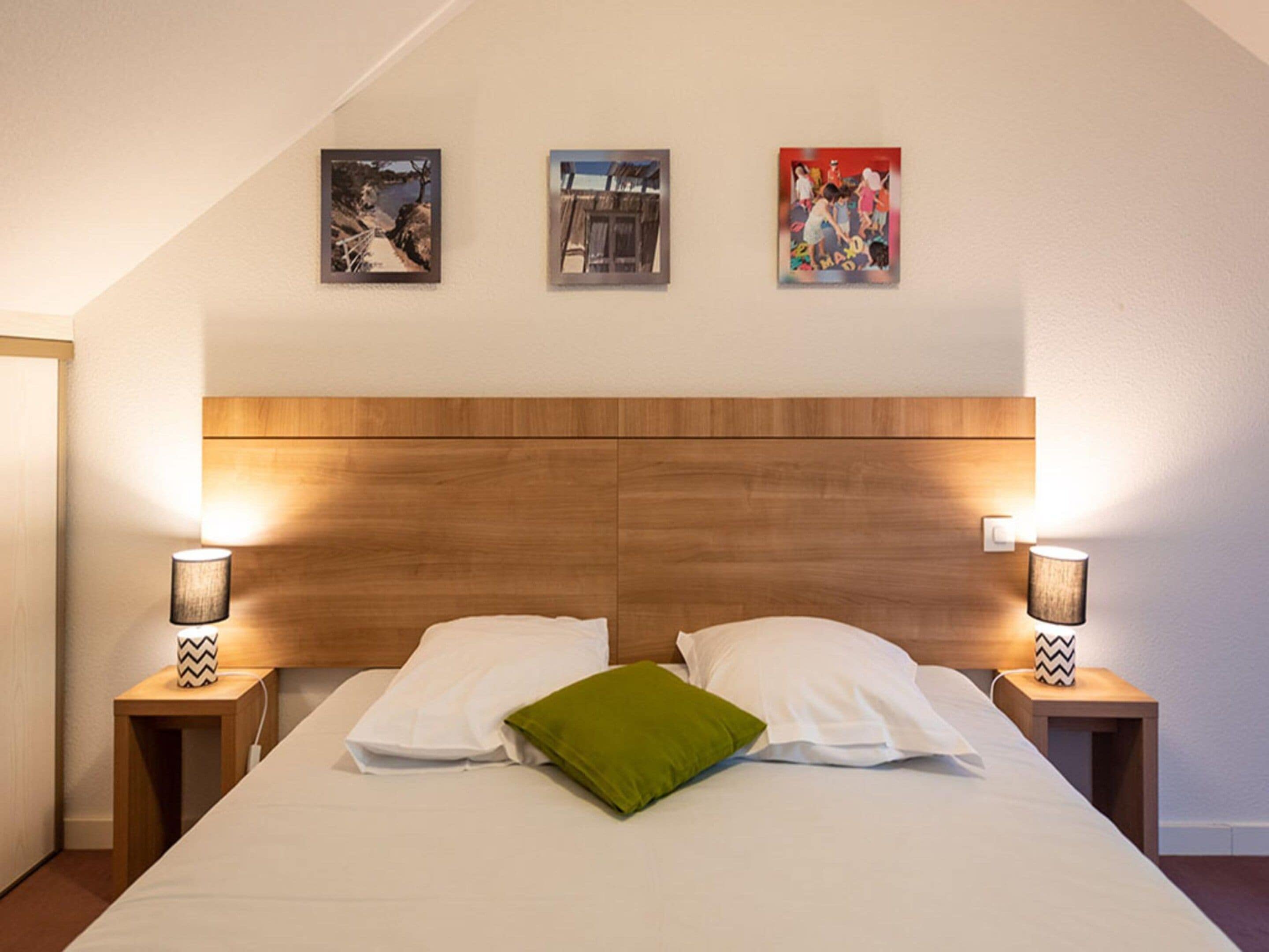 Une chambre à coucher avec une tête de lit en bois, deux lampes de chevet, un lit avec du linge de lit blanc et un oreiller vert, et trois tableaux encadrés accrochés au mur.