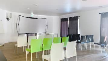A conference room with a white table, green and white chairs, a white screen, and a window with curtains.