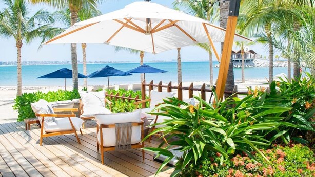 Un patio frente a la playa con sillas de madera, sombrillas blancas y abundante vegetación.