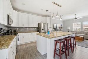 A modern kitchen with white cabinets, granite countertops, a central island with stools, stainless steel appliances, pendant lighting, and a ceiling fan. - Casa Rio - Canal Front, walk to the beach! (St. Augustine)