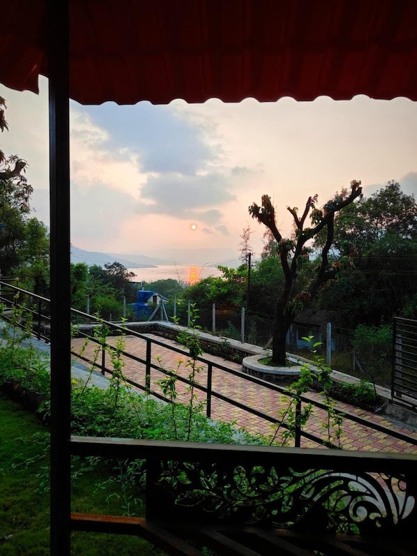 A view from a covered balcony with a metal railing, a paved walkway, and a tree in the foreground.