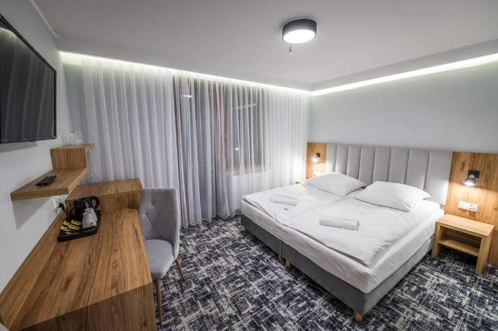 Una habitación de hotel con cama, escritorio, silla, televisor y ventana con cortinas.