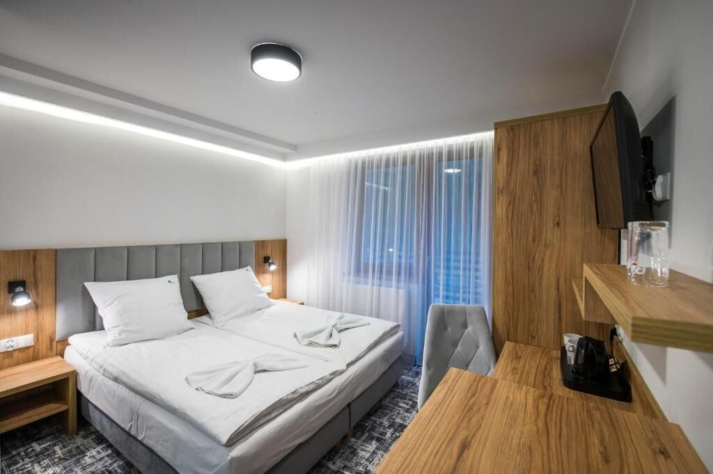 A hotel room with a bed, bedside tables, a desk, a chair, a TV, and a window with sheer curtains.