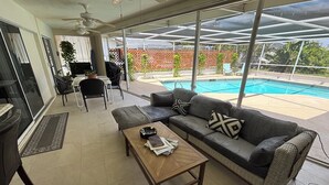 Uma sala de estar moderna com sofá, mesa de centro e cadeiras, uma porta de vidro que dá para o exterior, e vista para uma piscina.