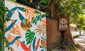 Un mural colorido en una pared, un letrero que dice 'SOLAR DAS PETALAS' y un árbol con una cerca al fondo.