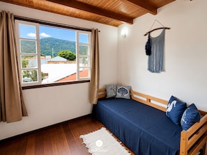 Una habitación con cama, ventana con cortinas, un macramé colgado en la pared y techo de madera.