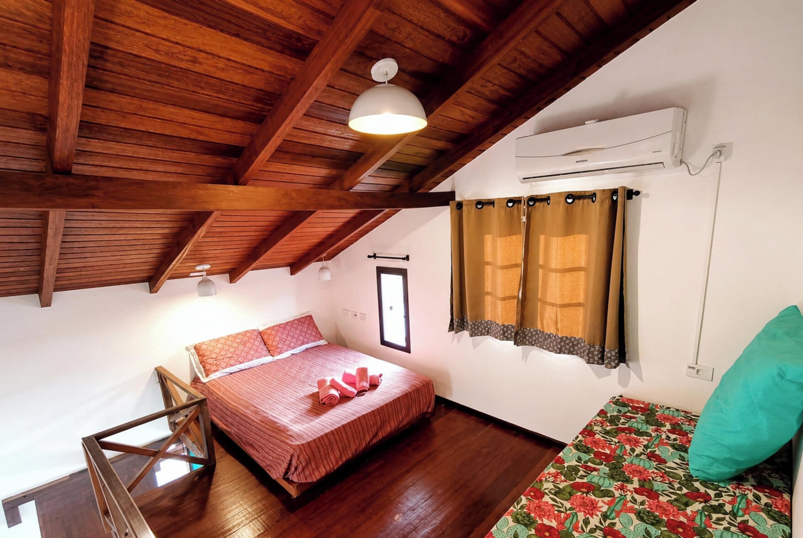 A bedroom with a wooden ceiling, a bed with red bedding, a window with curtains, and an air conditioning unit.