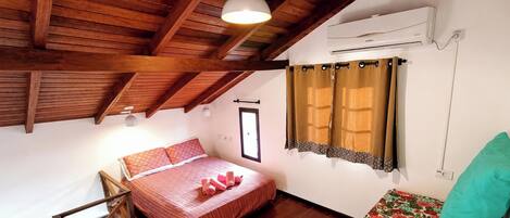 A bedroom with a wooden ceiling, a bed with red bedding, a window with curtains, and an air conditioning unit.
