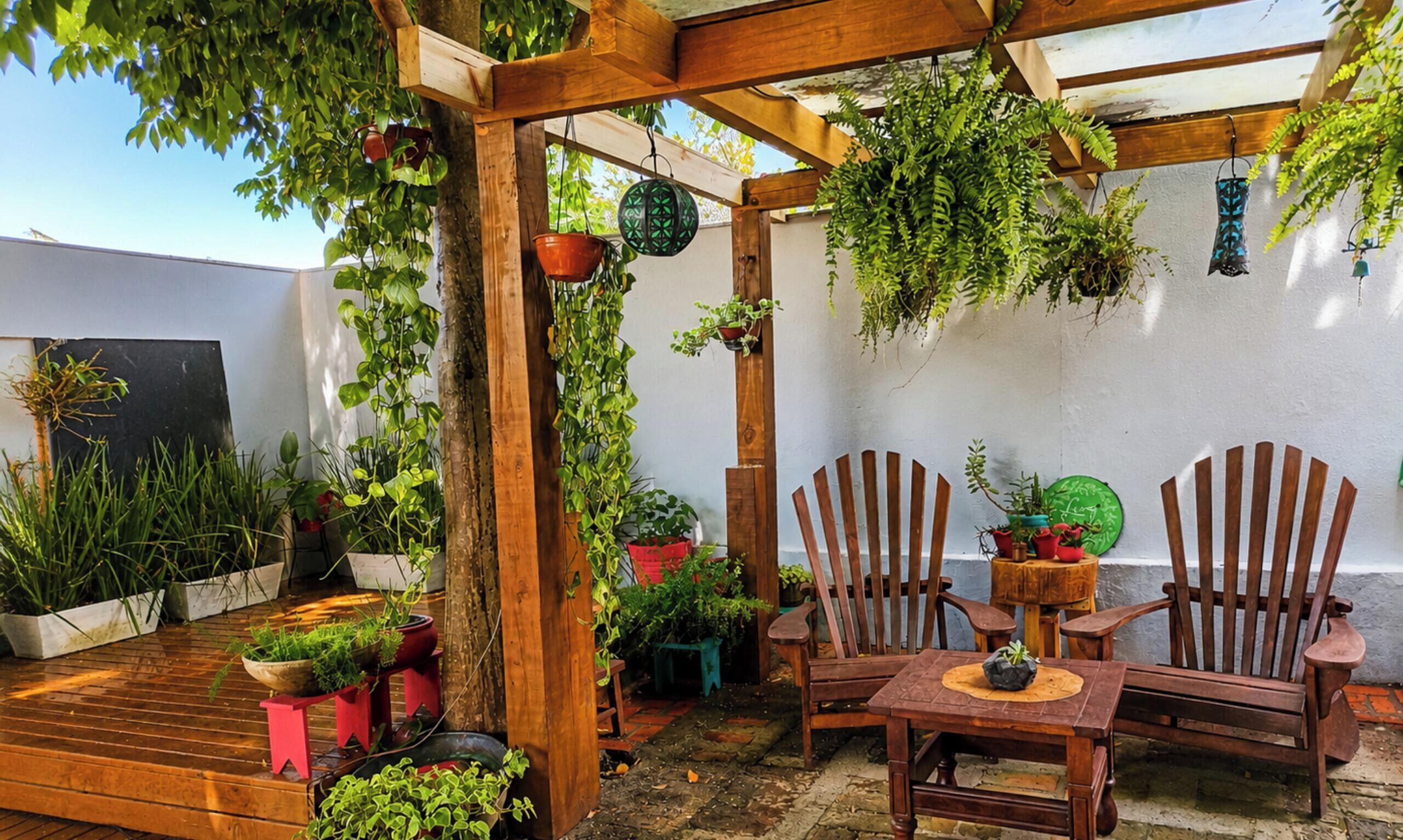 Una pérgola de madera con plantas colgantes, una mesa de madera con sillas y macetas con plantas en un patio.