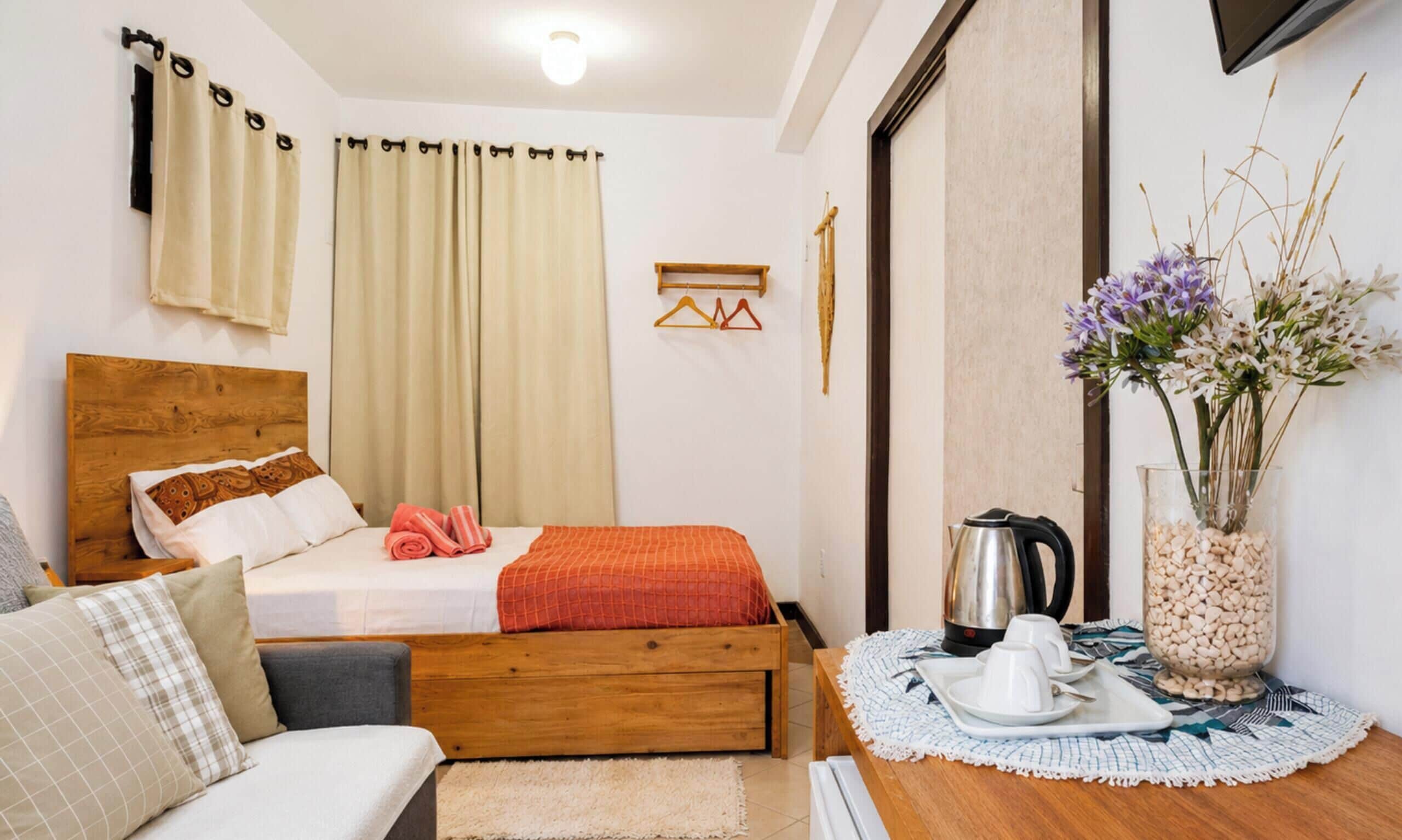 Une chambre à coucher avec un lit, une tête de lit en bois, une table de chevet avec une bouilloire, un vase avec des fleurs et un téléviseur fixé au mur.