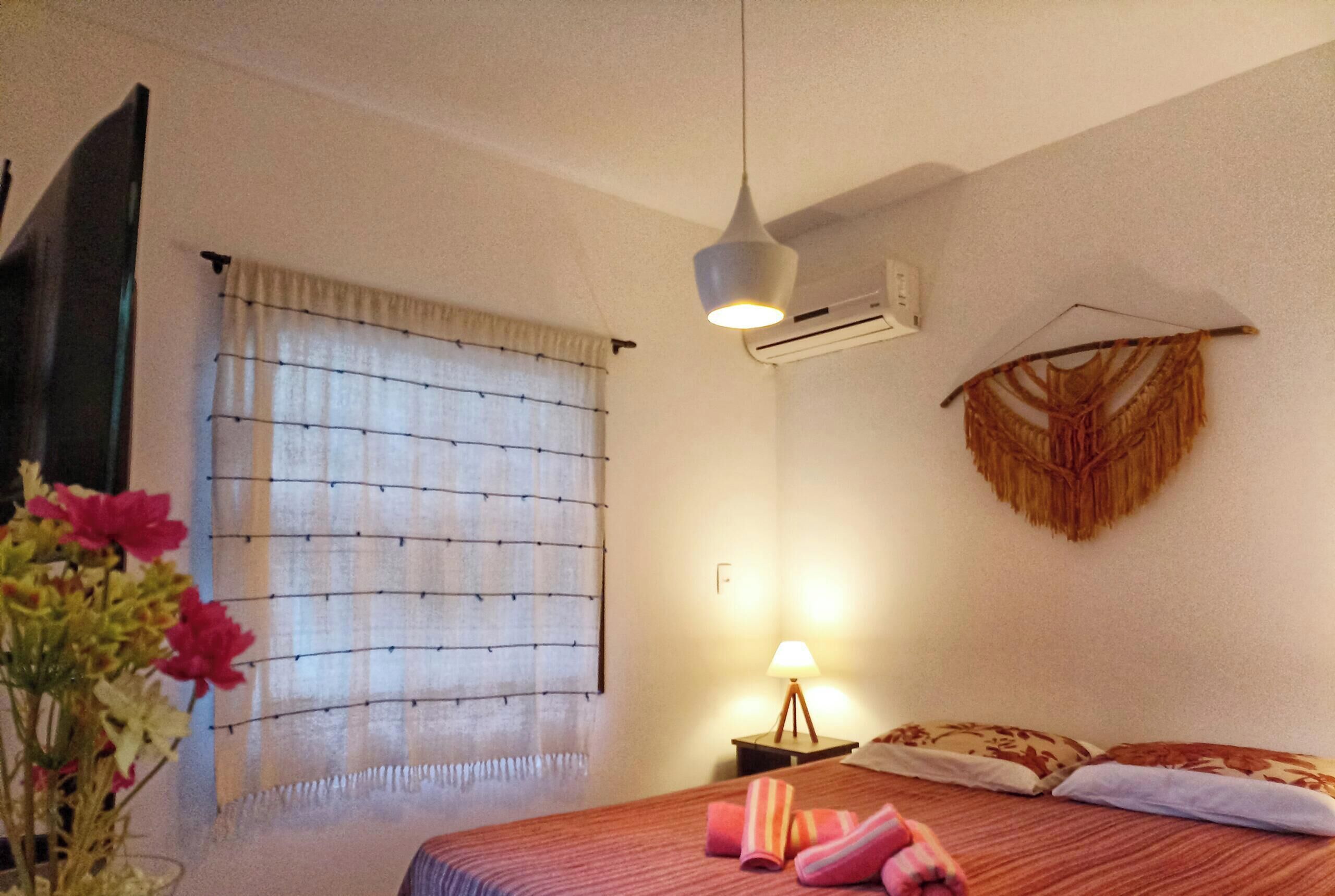 A hotel room with a bed, a bedside lamp, an air conditioning unit, a wall-mounted decorative item, and a window with curtains.