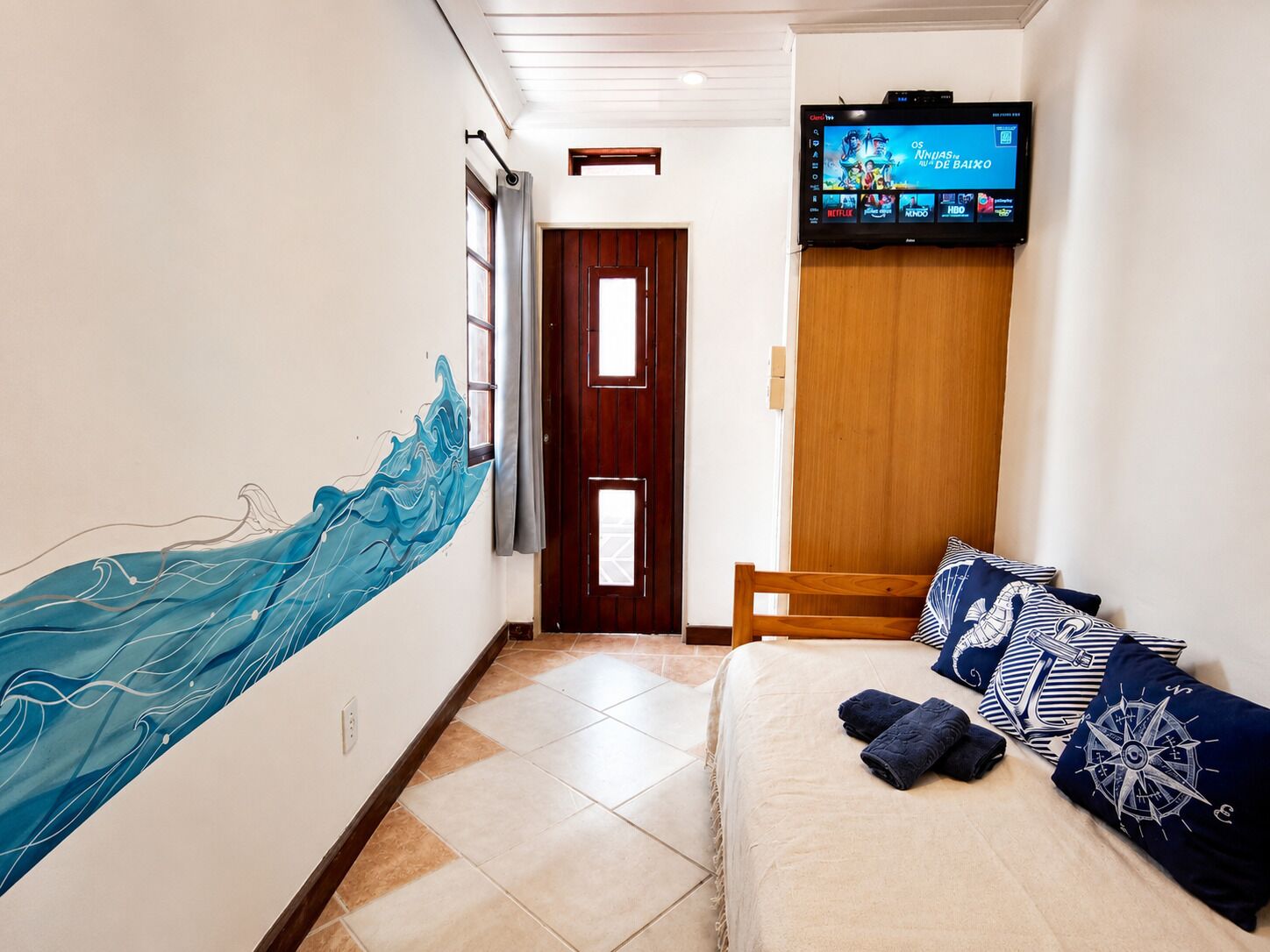 Ett hotellrum med en tv som är monterad på väggen, en säng med kuddar i marint tema och en stor, abstrakt målning på väggen.