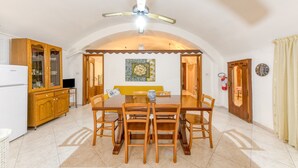 Un coin repas avec une table et des chaises en bois, un ventilateur de plafond, et une cuisine équipée de placards et d’un réfrigérateur.
