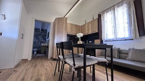 Moderni keittiö, jossa on puiset kaapit, ruokailutila pöytineen ja tuoleineen sekä olohuone, jossa on sohva.