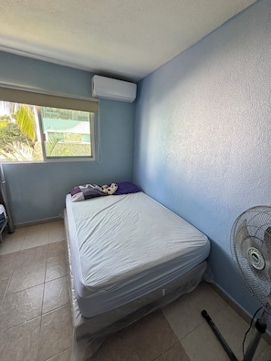 Una habitación con cama, ventana, aire acondicionado, ventilador y vistas a zonas verdes.