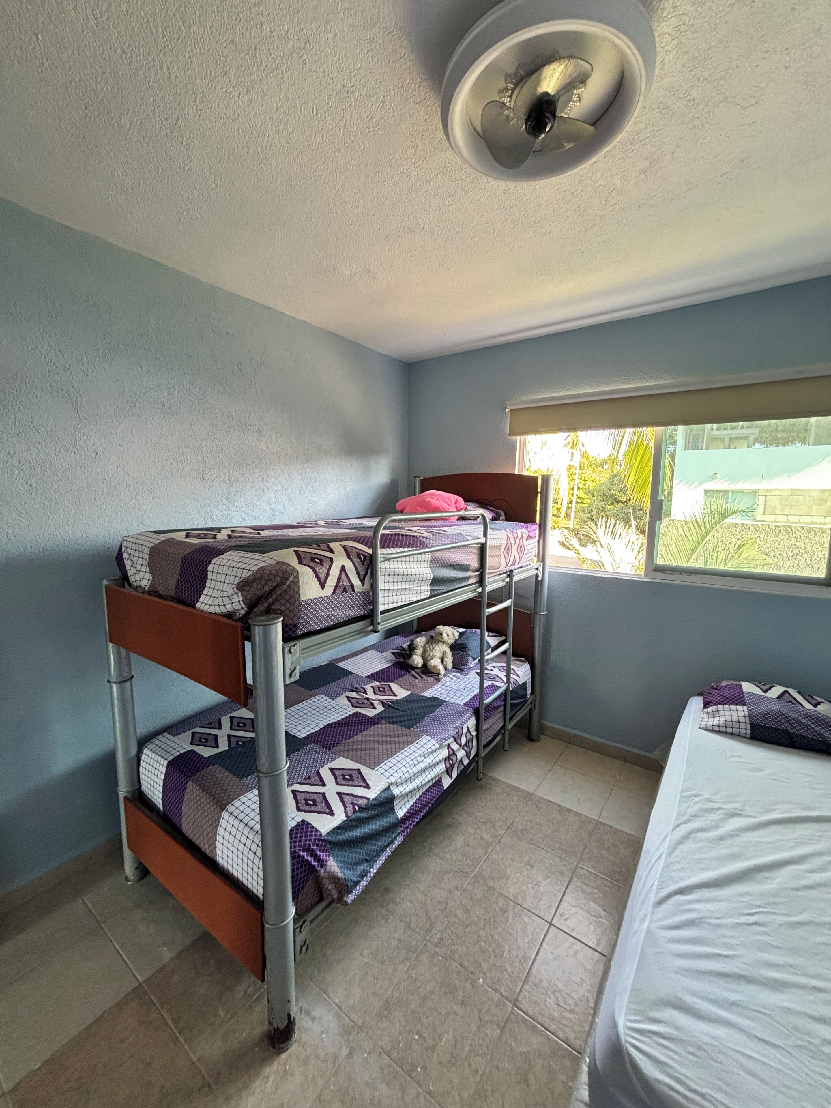 A bunk bed room with a window, a ceiling light, and a bed with a white mattress.