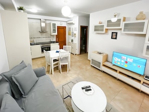A modern living room with a grey sofa, a dining table with chairs, a TV, and a kitchen in the background. - Holiday apartment in Mogan (Lomo Quiebre)
