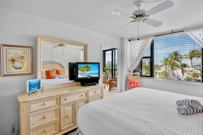 Habitación de hotel con cama, televisión, cómoda, espejo y ventana con vista a palmeras.