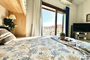 A hotel room with a bed, a TV, a window with curtains, and a plant on the windowsill. - MONO SUITE - Rena Bianca (Santa Teresa Gallura)