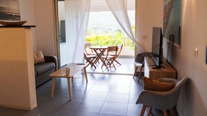Un espacio de sala con un sofá, una mesa de comedor con sillas, un televisor y vistas a zonas verdes por una ventana.