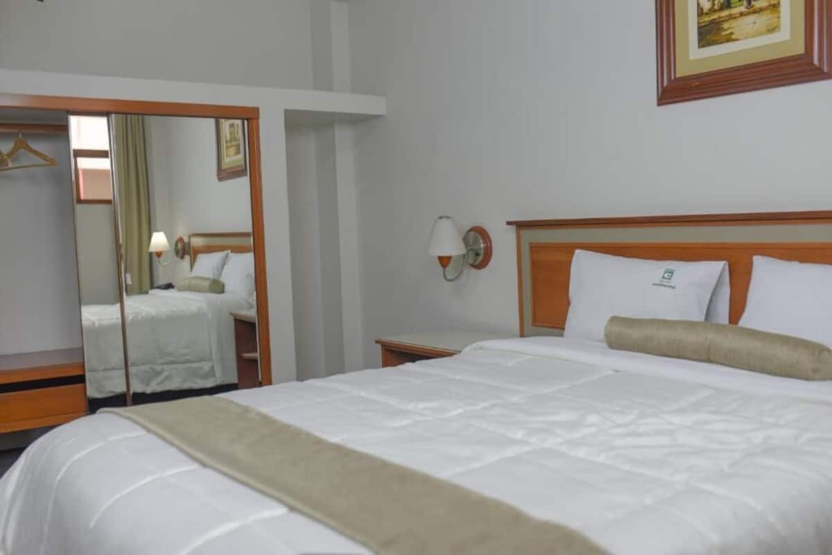 Habitación de hotel con cama, mesita de noche, espejo y lámpara de pared.