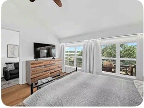 A bedroom with a bed, a TV, a wooden dresser, and large windows with a view. - The Sunset Deck - Lake Travis Beach House· Pool · Golf · Sleeps 12 (Lago Vista)