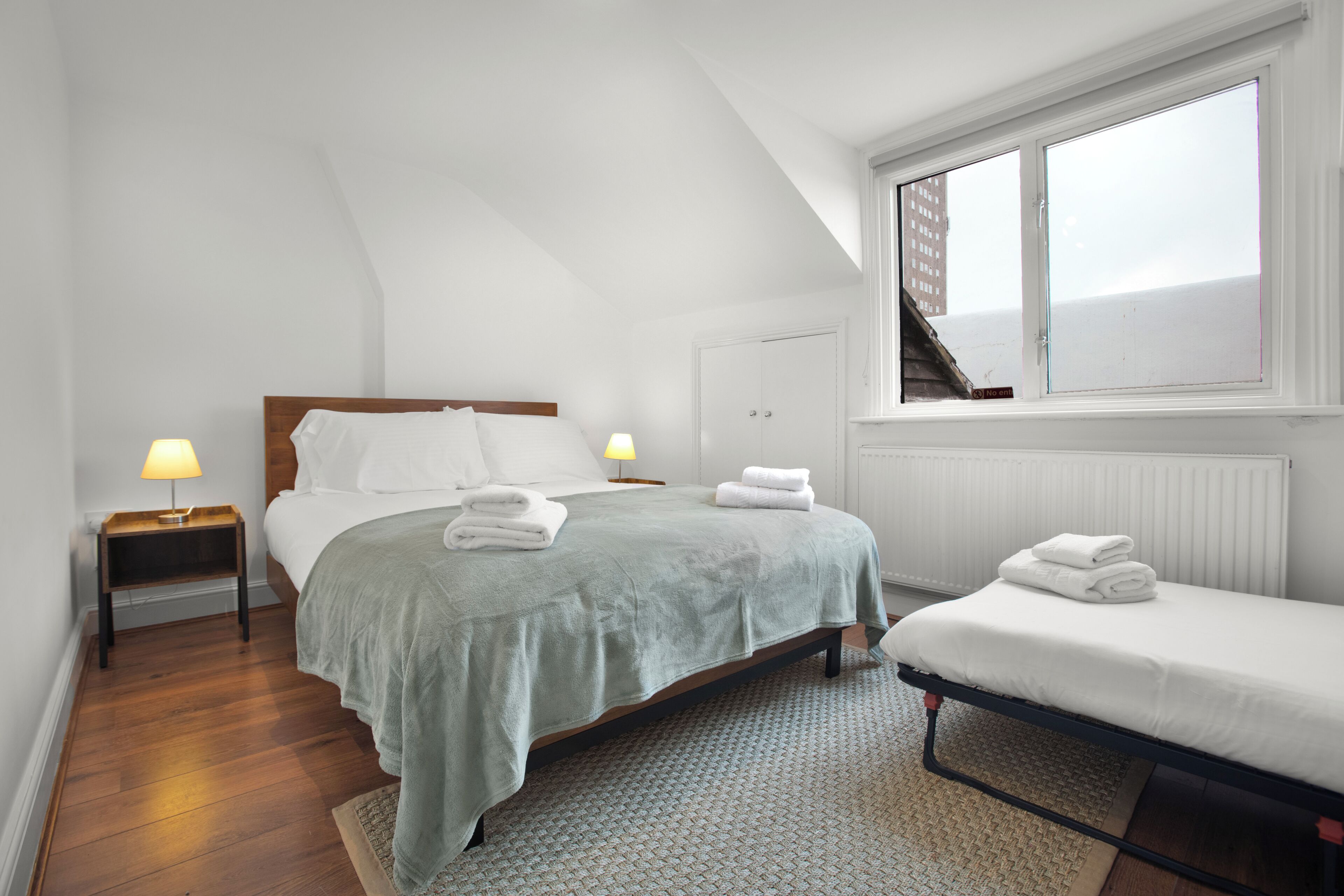 Une chambre à coucher avec un lit, une table de chevet, une fenêtre et une vue sur un bâtiment à l’extérieur.