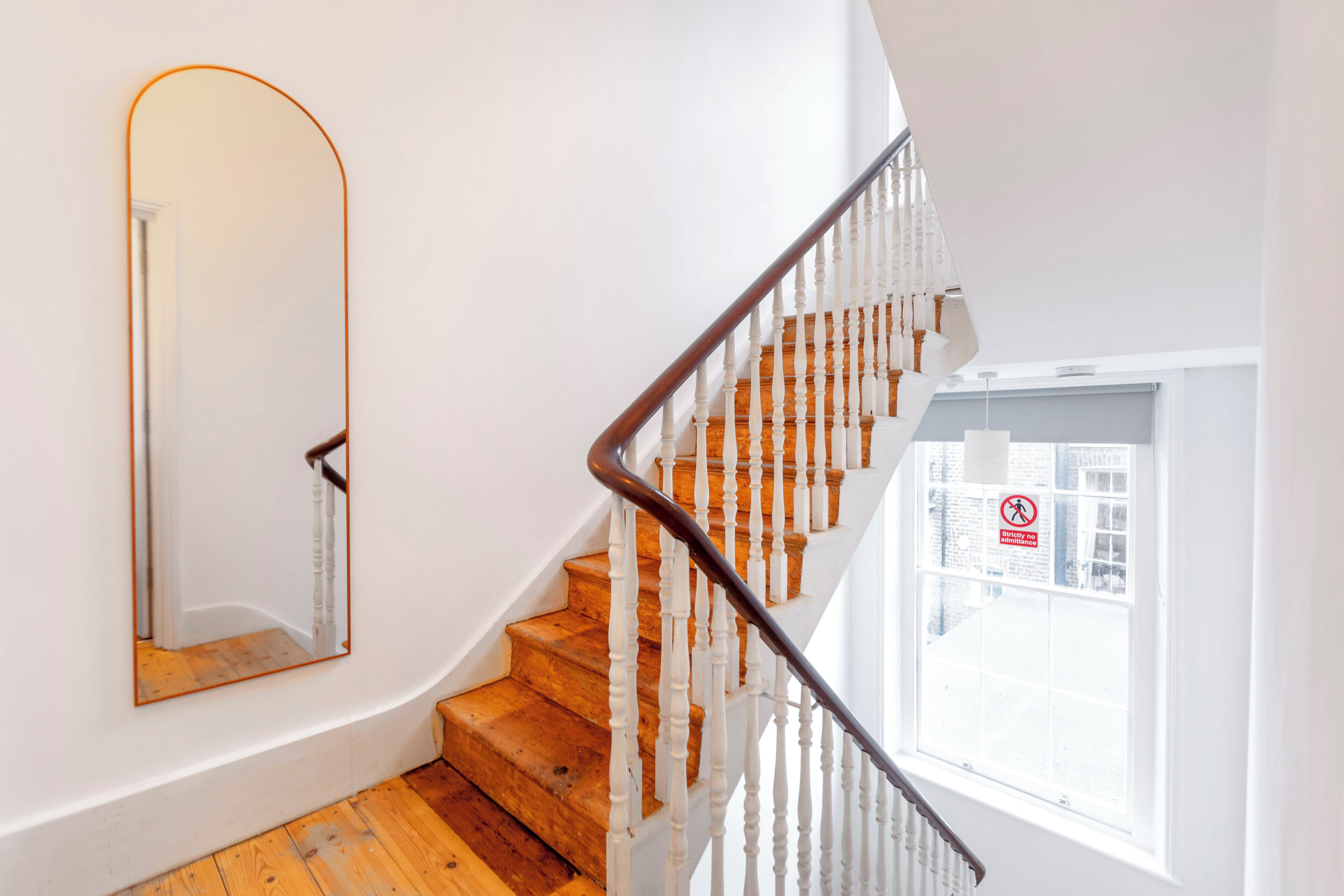 A staircase with wooden steps and white balusters, a mirror on the wall, and a window with a view of buildings.
