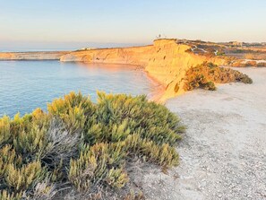 A coastal landscape with a sandy beach, rocky cliffs, and sparse vegetation. - Modern & Bright Maisonette • Private Terrace • Close to the Sea and Centre (Wied il-Għajn)