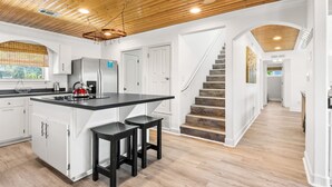 A modern kitchen with a central island, wooden ceiling, and a staircase leading to a second floor. - 🏝️Private Pool + Game Room | Sleeps 15 | Steps to Beach | OWB Beach House (Pensacola Beach)