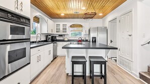 A modern kitchen with wooden ceiling, stainless steel appliances, white cabinetry, and a central island with stools. - 🏝️Private Pool + Game Room | Sleeps 15 | Steps to Beach | OWB Beach House (Pensacola Beach)
