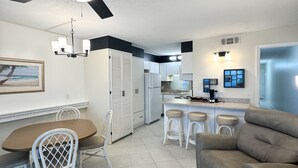 Cozinha moderna com armários brancos, sala de jantar com cadeiras de vime, ventilador de teto e um sofá cinzento.