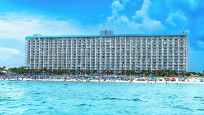 Um hotel grande com vários andares e varandas, uma praia com guarda-sóis e pessoas, e um céu azul e límpido.