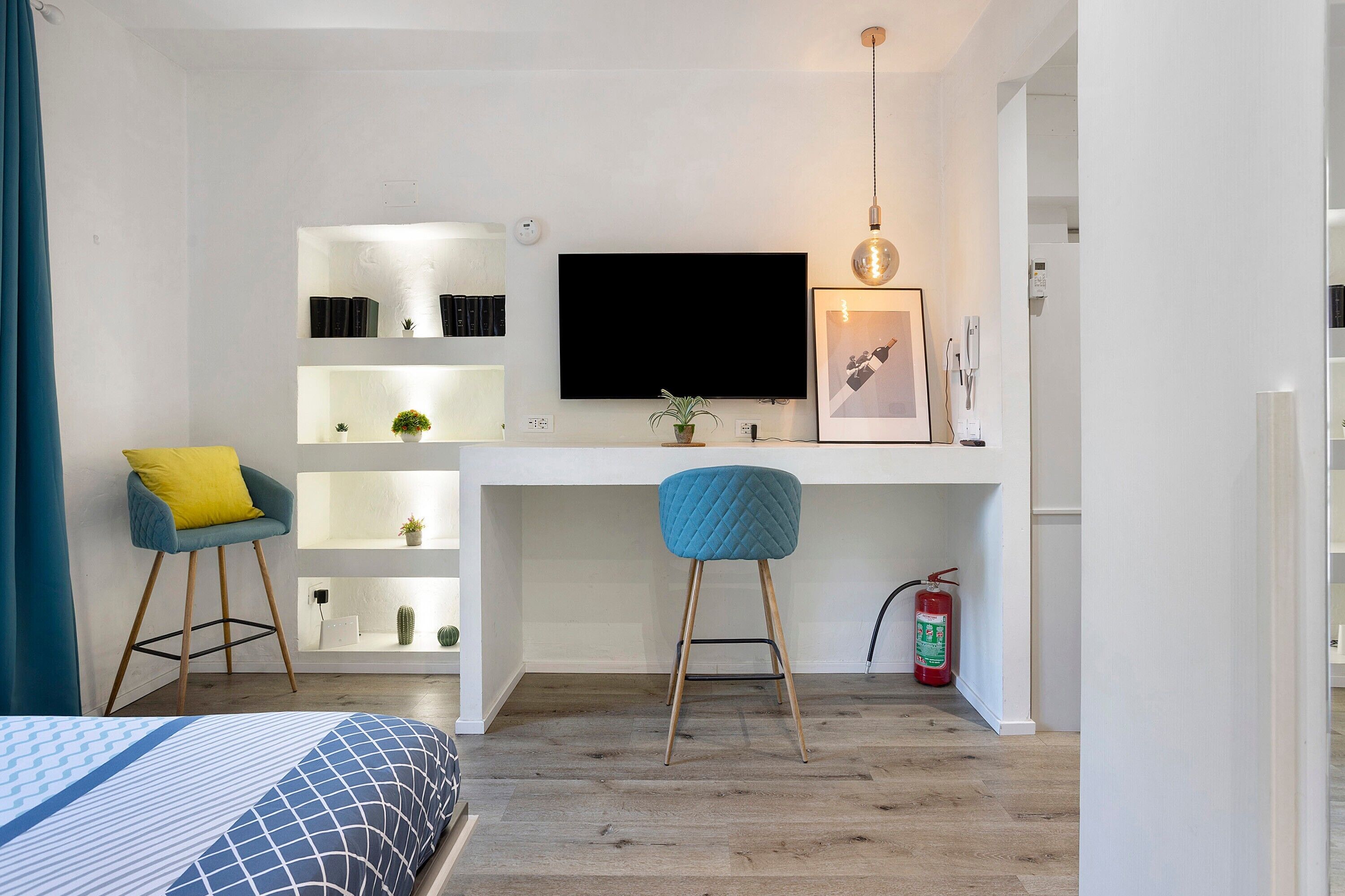 A modern bedroom with a bed, a desk with a TV, two chairs, a bookshelf, and a fire extinguisher.