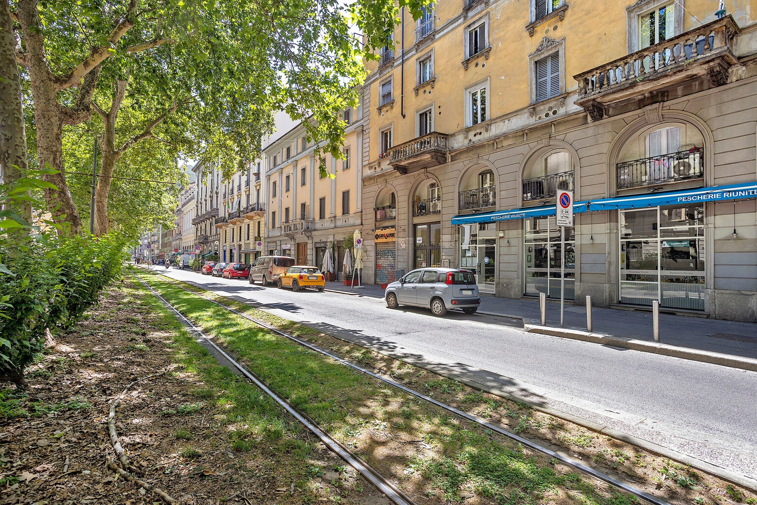 Una calle con edificios, una línea de tranvía y un toldo azul con el texto 'PESCHERIE RIUNITTE'.