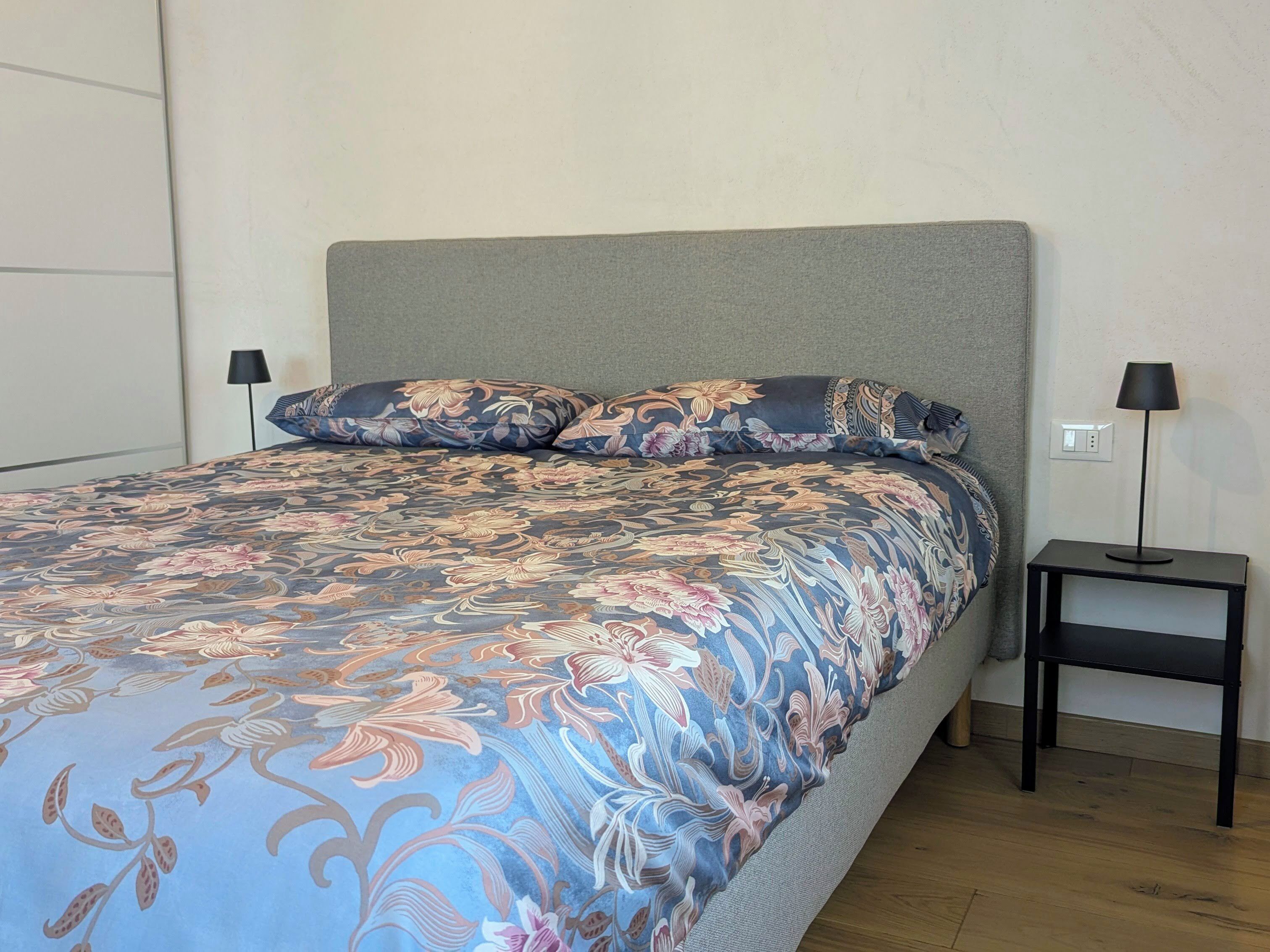 Ein Bett mit Blumenmuster auf der Bettwäsche, ein grauer Kopfteil und zwei Nachttische mit Lampen.