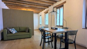A modern living room with a wooden ceiling, a green sofa, a dining table with chairs, and large windows.