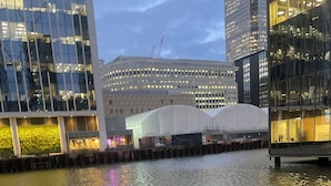 Blick auf das Wasser mit modernen Gebäuden, ein weißes Bauwerk mit gewölbtem Dach und ein Metallgeländer im Vordergrund.