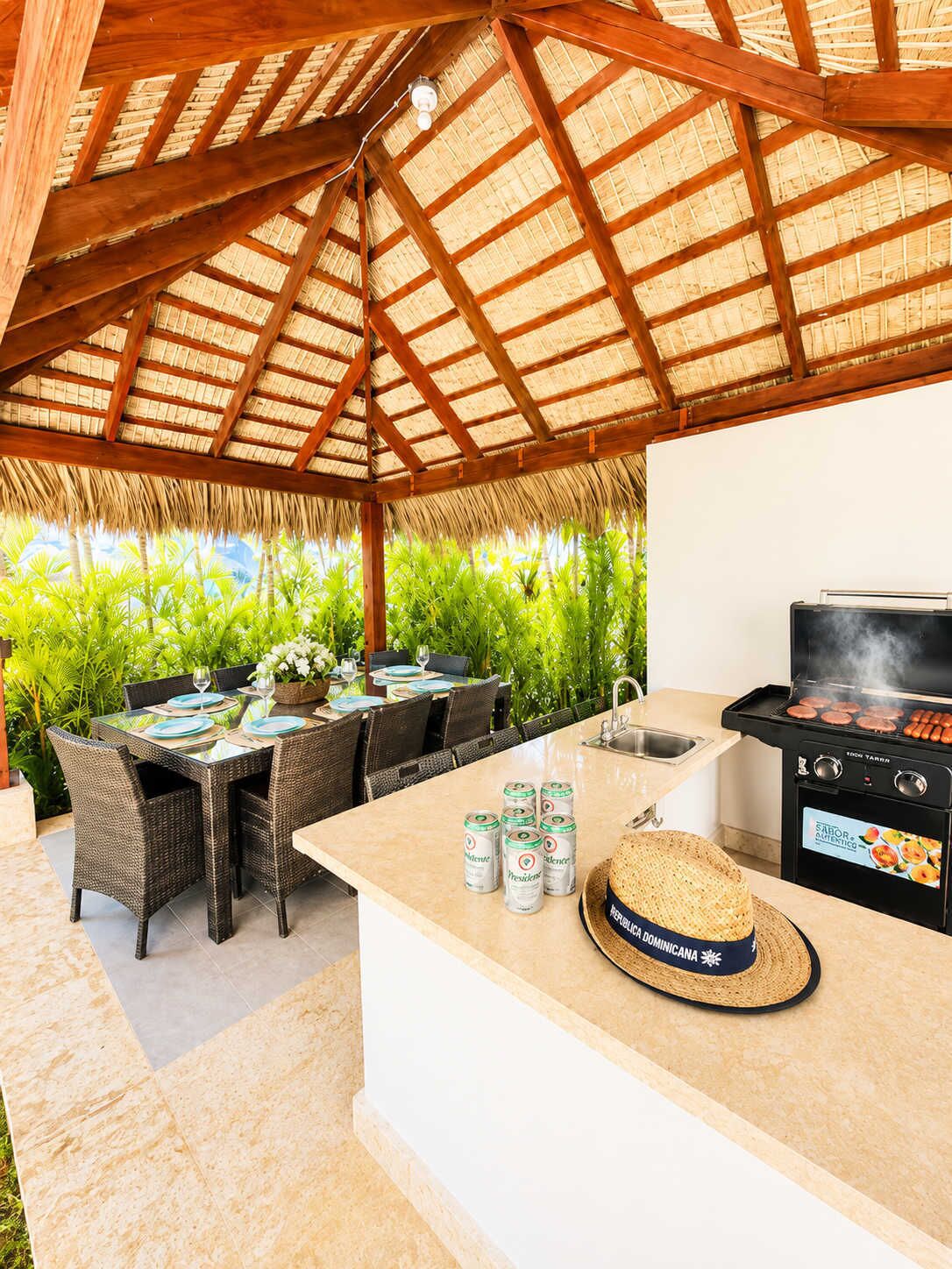 An outdoor dining area with a grill, wicker furniture, and a thatched roof.